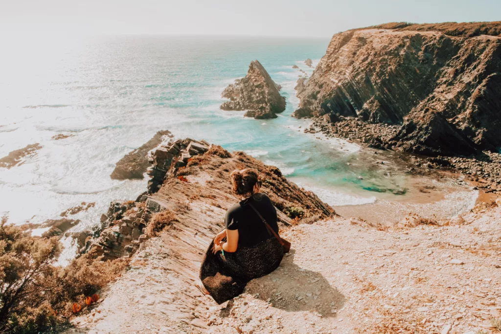 A traveler sitting on a cliff overlooking the rugged coastline of Portugal’s Alentejo region, highlighting slow travel and sustainable tourism.