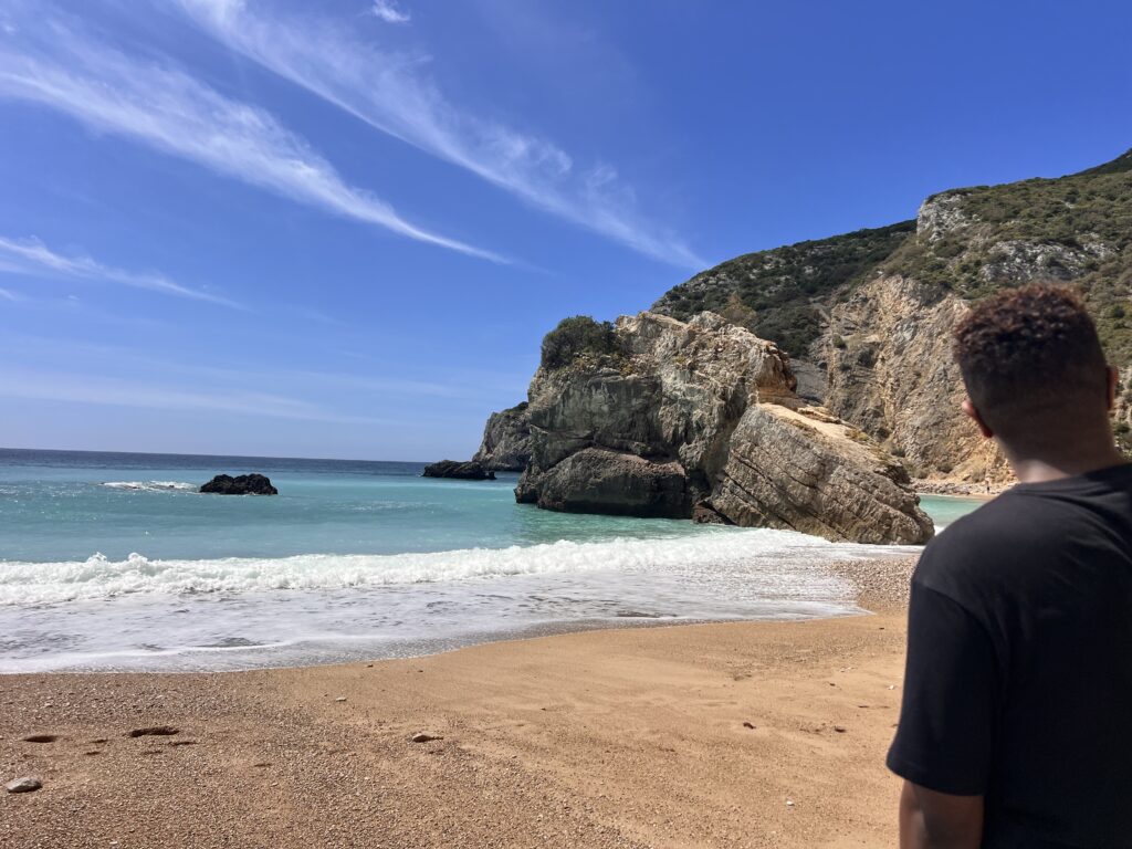 A person standing on the beach at Carvalho Beach near Sesimbra, Portugal, facing the ocean with cliffs rising around the shoreline — showcasing sustainable and slow coastal travel.